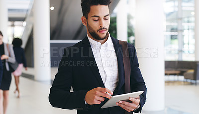 Buy stock photo Cropped shot of a handsome young businessman using a tablet while walking through the office during the day
