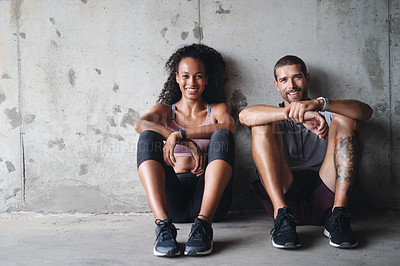 Buy stock photo Portrait of a sporty young couple sitting down against a wall while exercising inside a parking lot