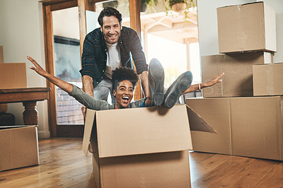 Buy stock photo Homeowners moving in, having fun, feeling carefree and excited while playing, joking and laughing. Young interracial couple relocating or buying a new property and sitting in a cardboard box at home