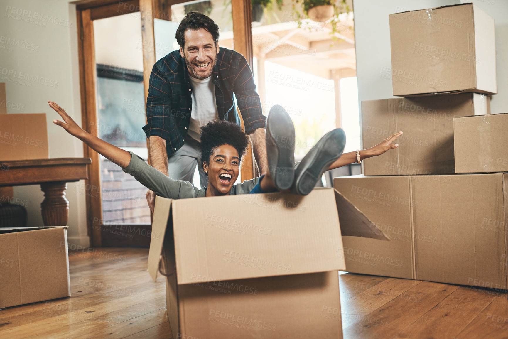 Buy stock photo Homeowners moving in, having fun, feeling carefree and excited while playing, joking and laughing. Young interracial couple relocating or buying a new property and sitting in a cardboard box at home