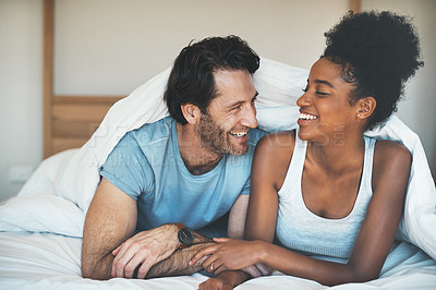 Buy stock photo Happy, carefree and laughing couple having fun lying in bed together. Interracial husband and wife bonding and showing affection while talking. Smiling lovers enjoying time indoors being playful 