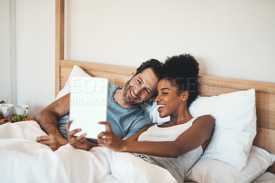Buy stock photo Happy couple taking selfie on a tablet in bed together after waking up. Interracial husband and wife enjoying their relationship, having fun while bonding and relaxing indoors on a weekend