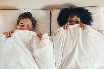 Buy stock photo High angle shot of a playful young couple covering themselves with blanket in bed at home