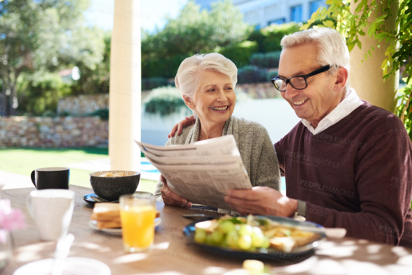 Buy stock photo Shot of an affectionate senior couple reading the newspaper while enjoying a meal together outdoors