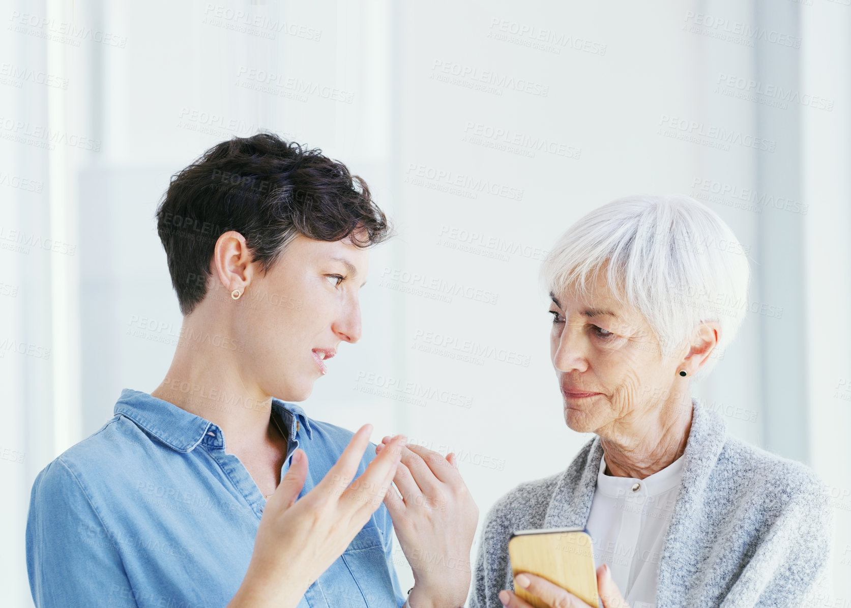 Buy stock photo Cropped shot of a senior woman holding a cellphone while talking with her attractive young daughter while at home