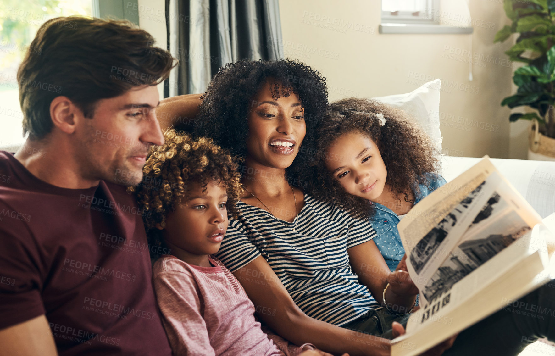 Buy stock photo Shot of a beautiful young family of four reading a story book together while relaxing on a sofa at home