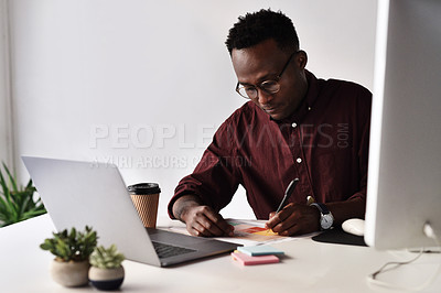 Buy stock photo Cropped shot of a handsome young businessman sitting alone and making notes while using his laptop in the office
