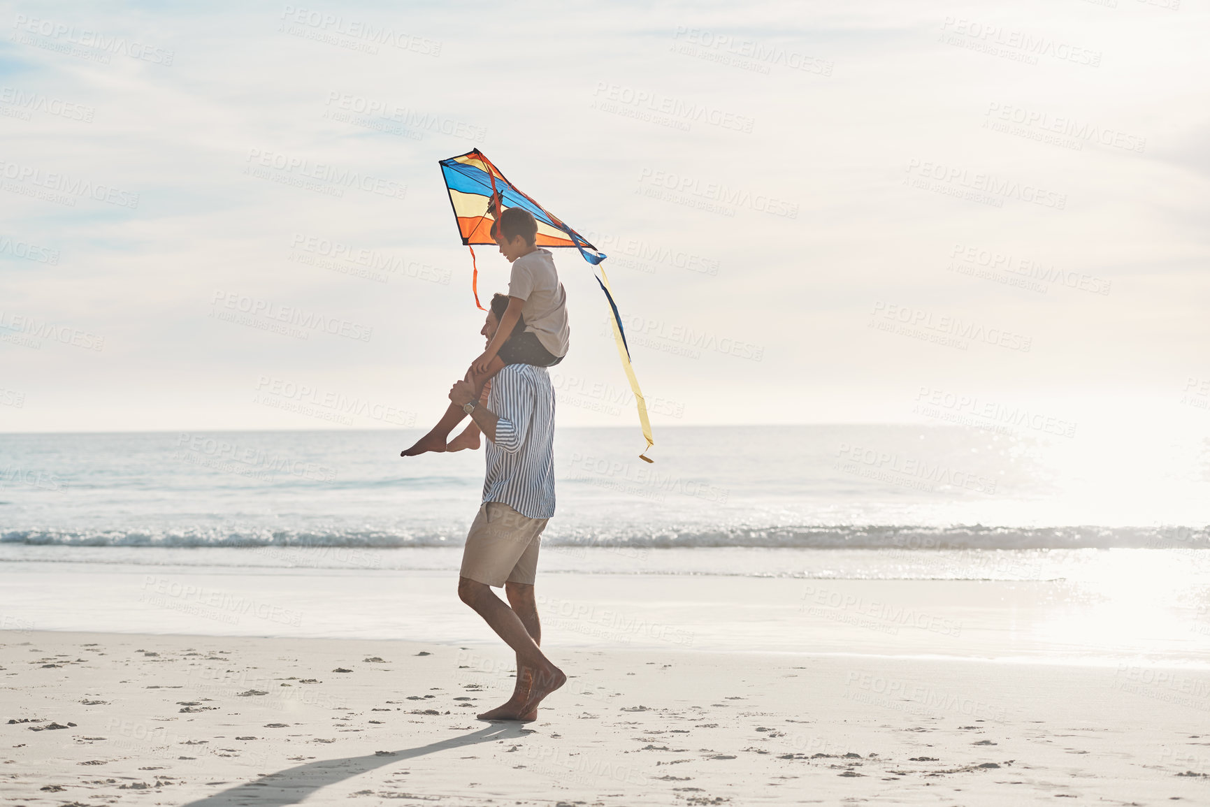 Buy stock photo Full length shot of a young boy being carried on his father's shoulders and holding a kite on the beach