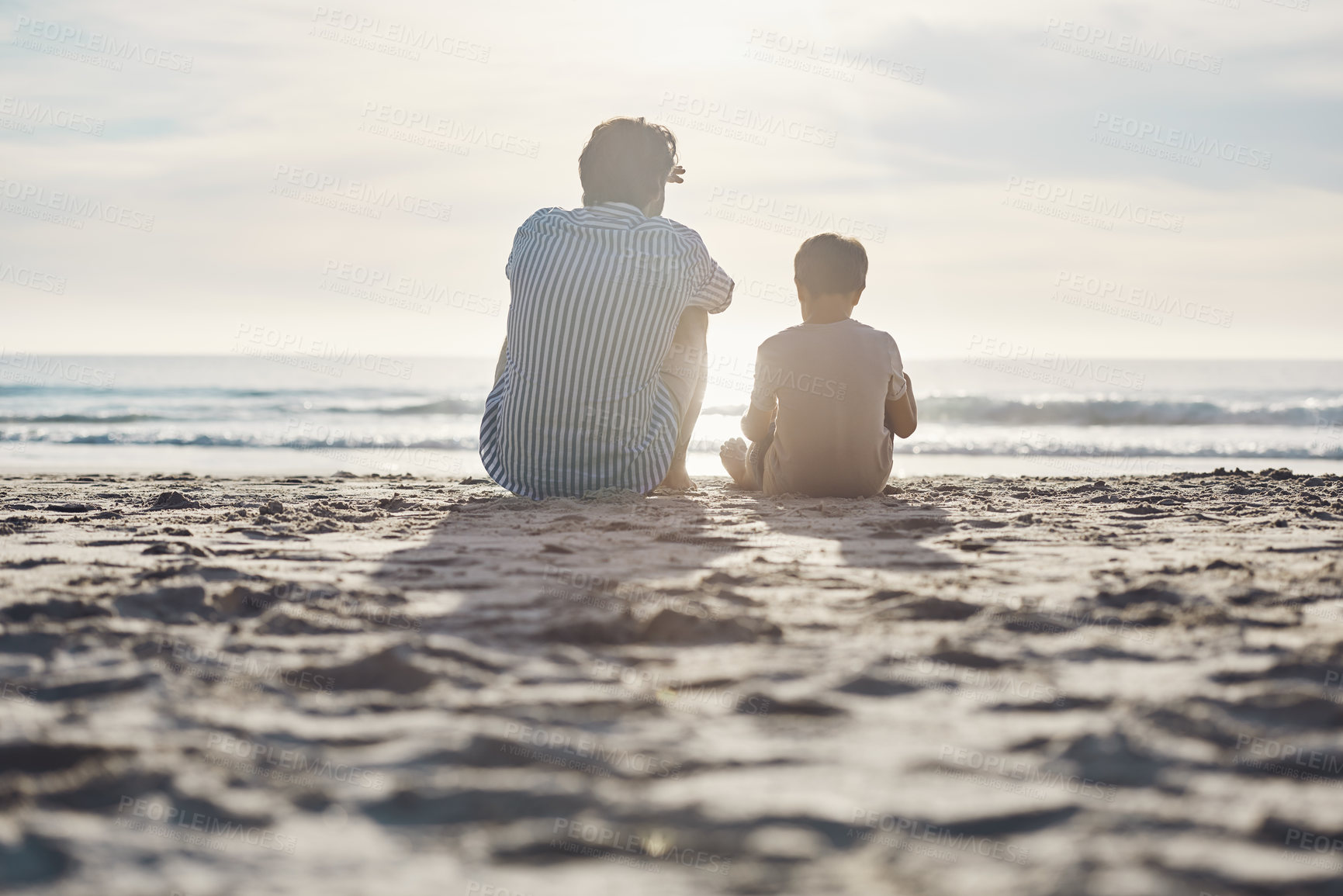 Buy stock photo Rearview shot of an unrecognizable father sitting and bonding with his son during an enjoyable day on the beach