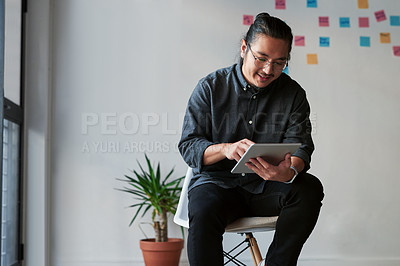 Buy stock photo Shot of a handsome young businessman sitting on a chair and using his digital tablet at work