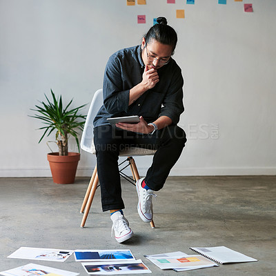 Buy stock photo Full length shot of a creative young businessman sitting on a chair and using his digital tablet at work