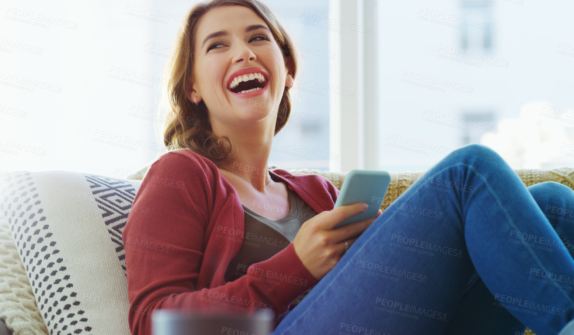 Buy stock photo Cropped shot of an attractive young woman sitting on her sofa alone and laughing while using her cellphone