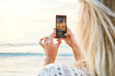 Buy stock photo Rearview shot of a woman taking a picture of the sun setting over the ocean