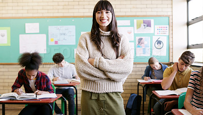 Buy stock photo Portrait of a cheerful young female teacher standing with her arms folded while her students do work inside of a classroom at school
