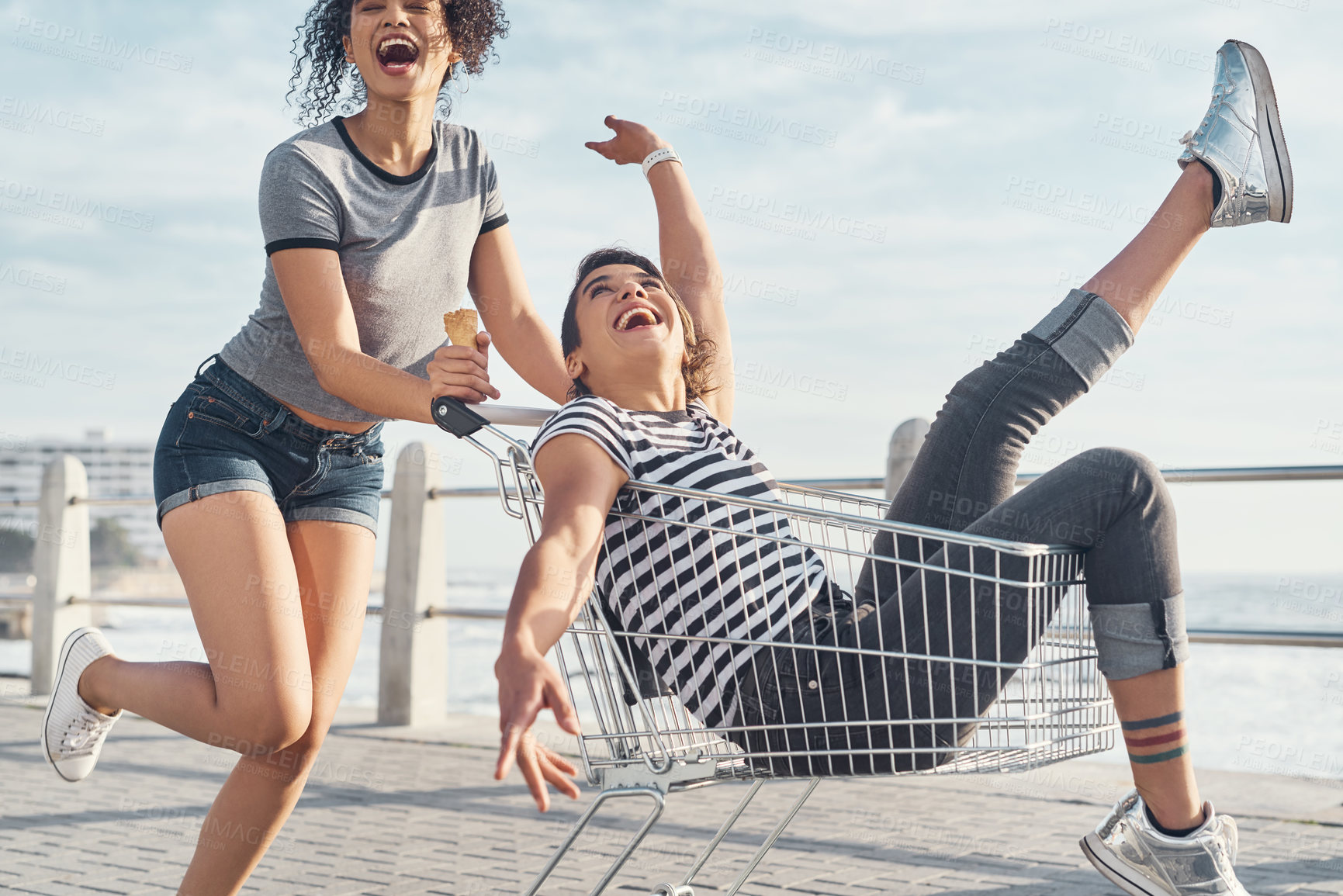 Buy stock photo Shot of a young woman pushing her friend around on the promenade in a shopping chart