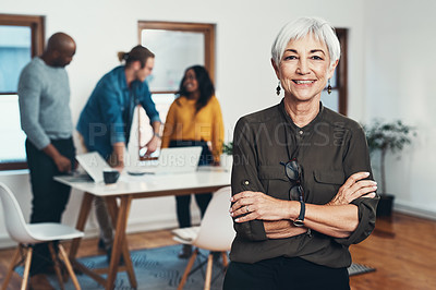 Buy stock photo Portrait of a cheerful mature businesswoman standing with her arms folded and her work colleagues in the background inside of the office