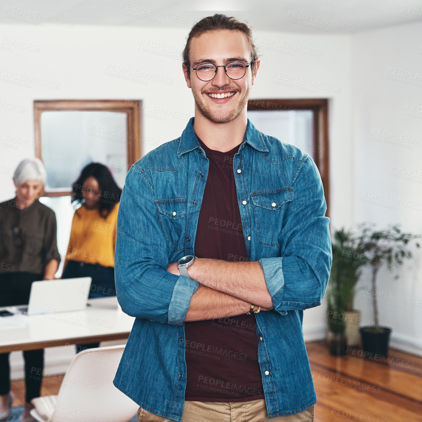 Buy stock photo Portrait of a cheerful young businessman standing with his arms folded and his work colleagues in the background inside of the office