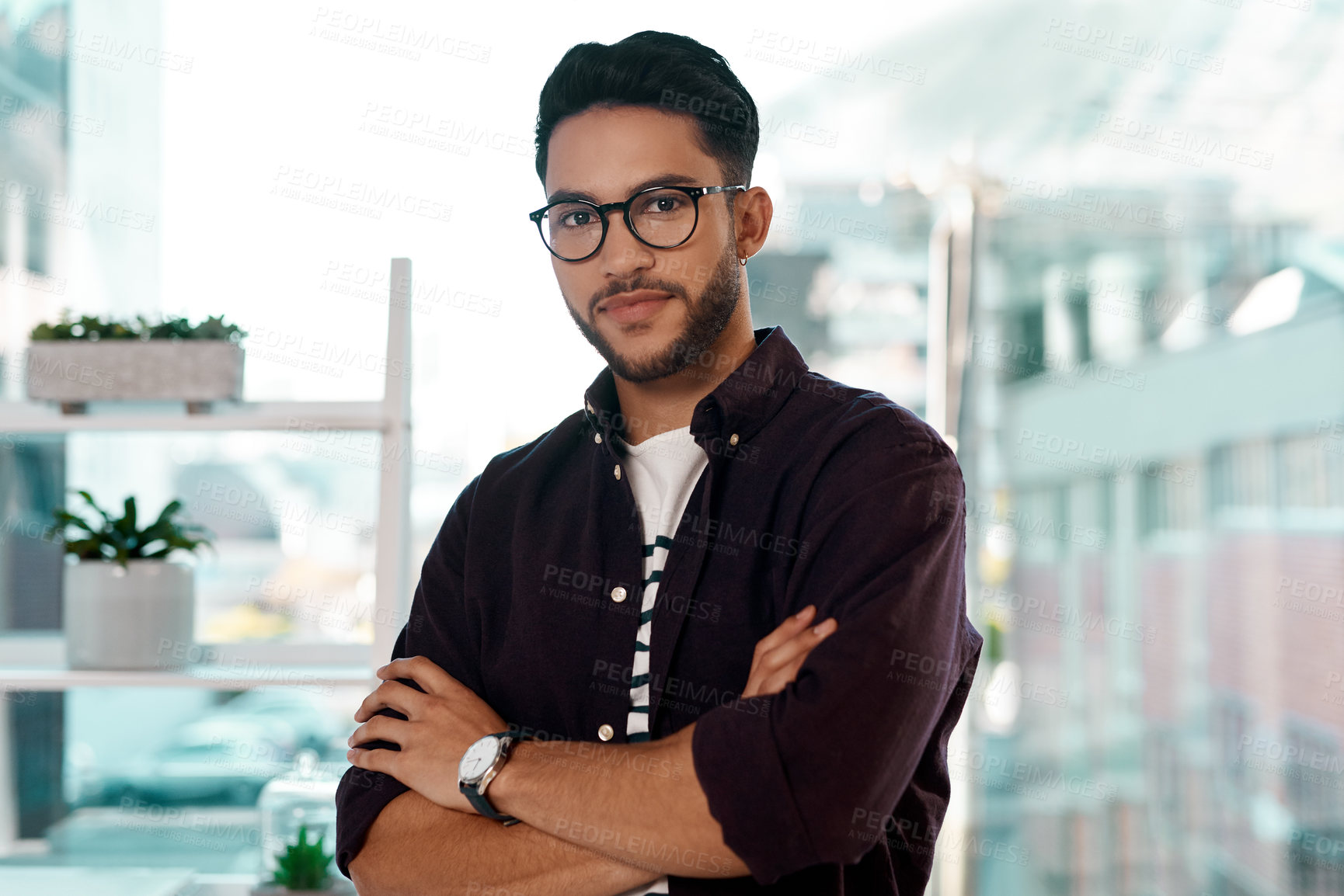 Buy stock photo Cropped portrait of a handsome young businessman wearing spectacles and standing alone in his office with his arms folded