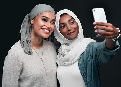Buy stock photo Cropped shot of two attractive young women standing together and wearing hijabs while taking a selfie against a black background