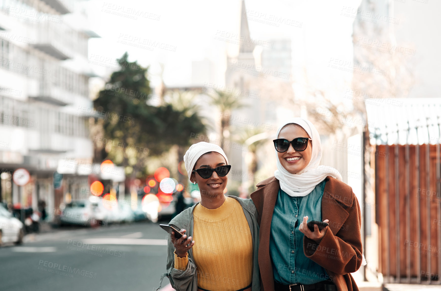 Buy stock photo Cropped shot of two attractive young muslim women wearing sunglasses and holding their cellphones while city sight-seeing