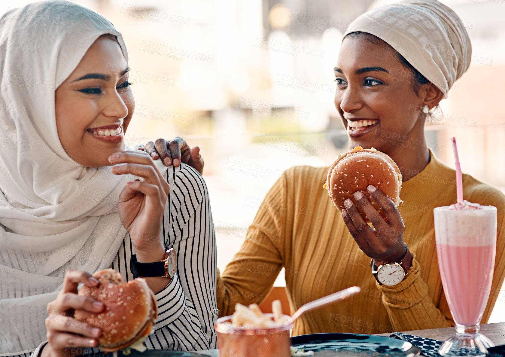 Buy stock photo Cropped shot of two affectionate young girlfriends eating burgers at a cafe while dressed in hijab
