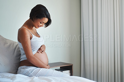 Buy stock photo Shot of a happy young pregnant woman sitting on a bed and holding her belly in her bedroom at home