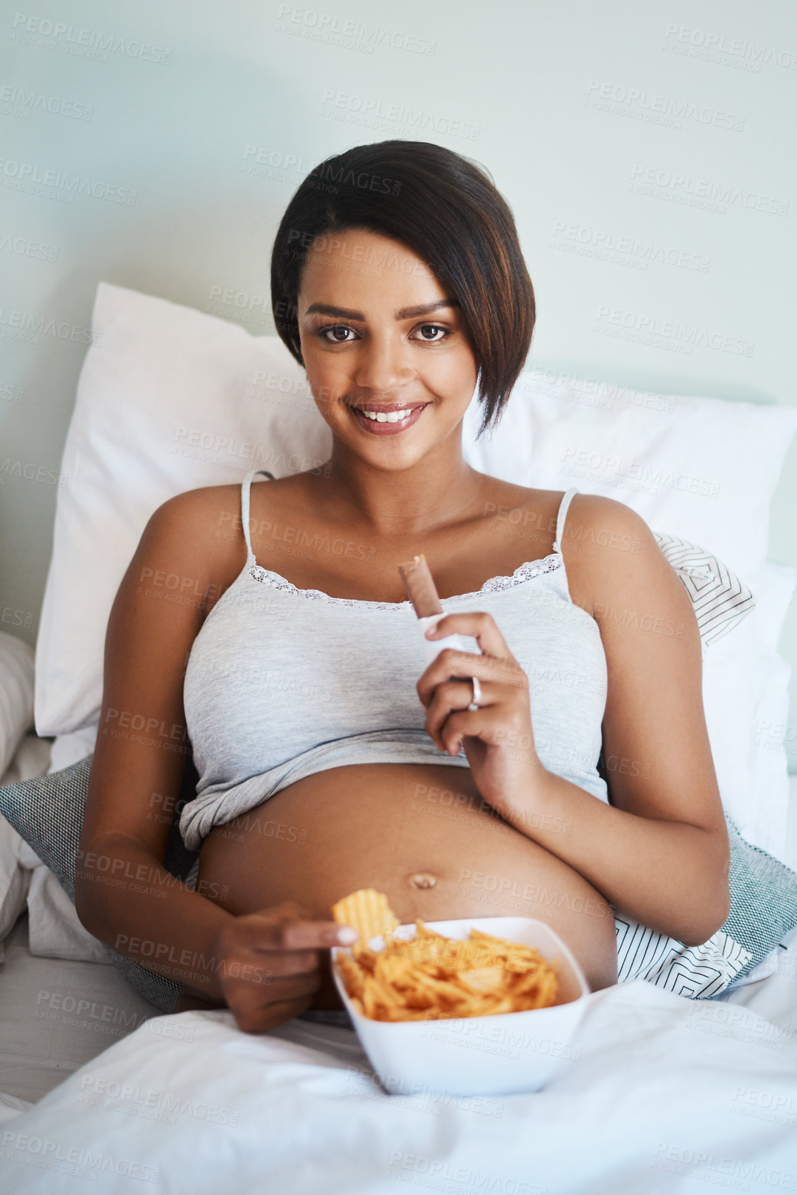 Buy stock photo Portrait of an attractive young pregnant woman eating potato chips and chocolate in bed