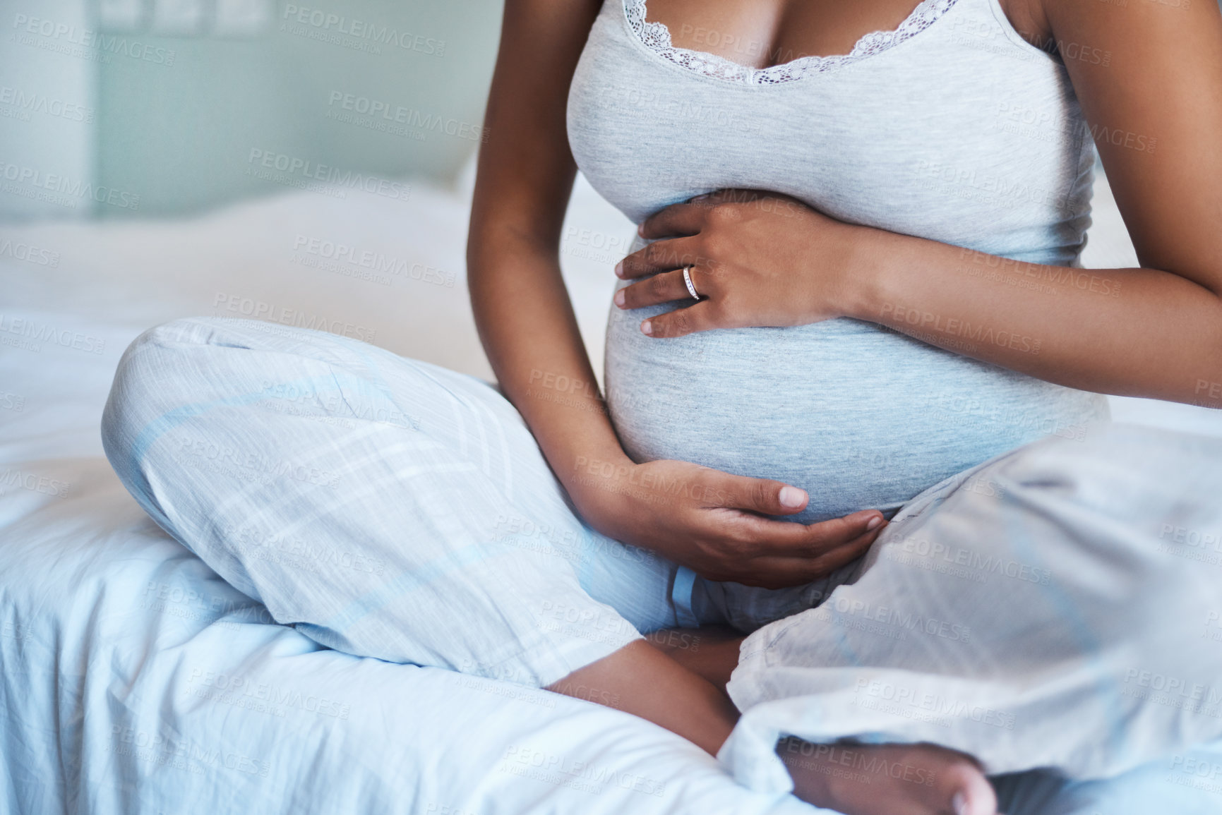 Buy stock photo Cropped shot of an unrecognizable pregnant woman sitting on a bed and holding her belly at home