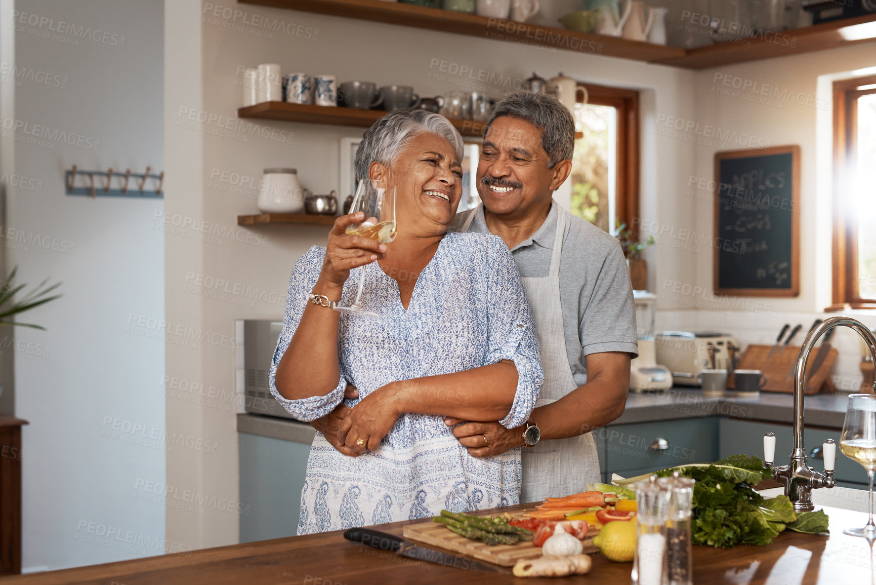 Buy stock photo Happiness, hug and old couple at kitchen counter with wine, food and cooking healthy dinner together. Smile, embrace and love, senior man and happy woman in retirement with drinks, vegetables and fun