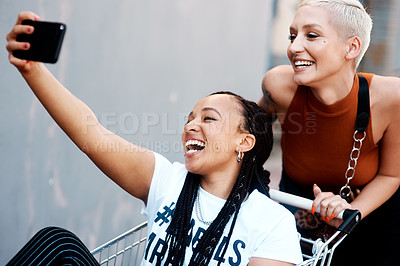 Buy stock photo Cropped shot of two cheerful young girlfriends taking a selfie while playing around with a shopping cart outdoors