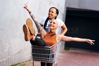 Buy stock photo Cropped shot of an energetic young woman pushing her female friend in a shopping cart outdoors
