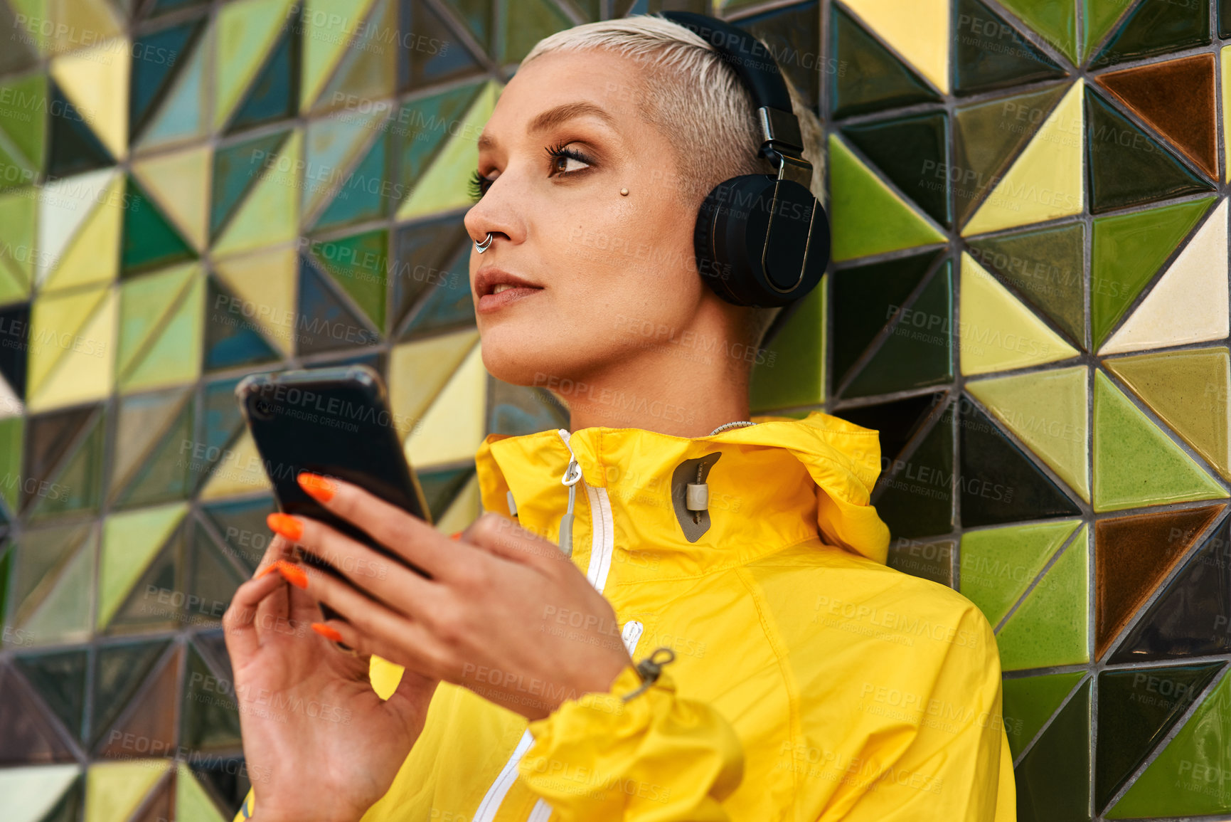 Buy stock photo Cropped shot of an attractive young woman standing against a wall and listening to music from her cellphone through headphones
