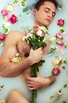 Buy stock photo High angle shot of a handsome young man holding a bouquet while lying in a bathtub full of milky water and flower petals at home