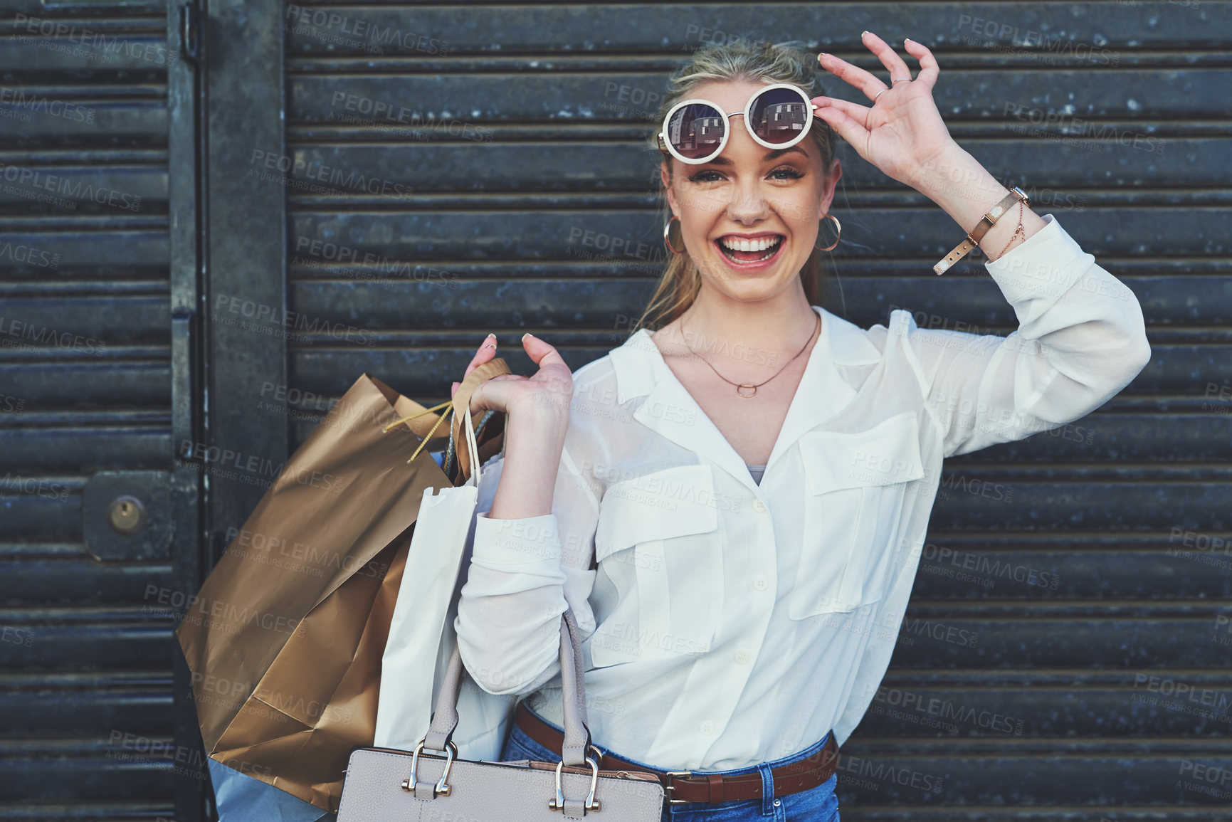 Buy stock photo Cropped portrait of an attractive young woman lifting her sunglasses while holding shopping bags against an urban background
