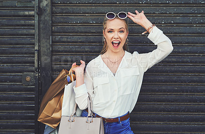 Buy stock photo Cropped portrait of an attractive young woman lifting her sunglasses while holding shopping bags against an urban background