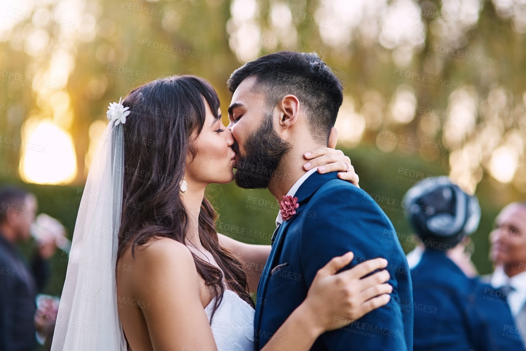 Buy stock photo Cropped shot of an affectionate young newlywed couple kissing passionately on their wedding day with their guests in the background
