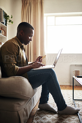 Buy stock photo Full length shot of a handsome young man using a laptop and a credit card to shop online while sitting on his couch at home