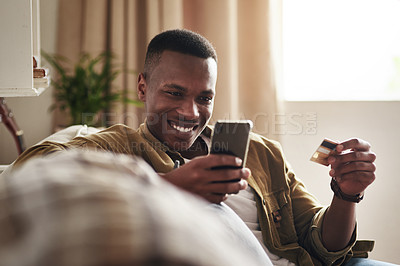 Buy stock photo Cropped shot of a handsome young man smiling while using a smartphone and a credit card to shop online in his living room at home