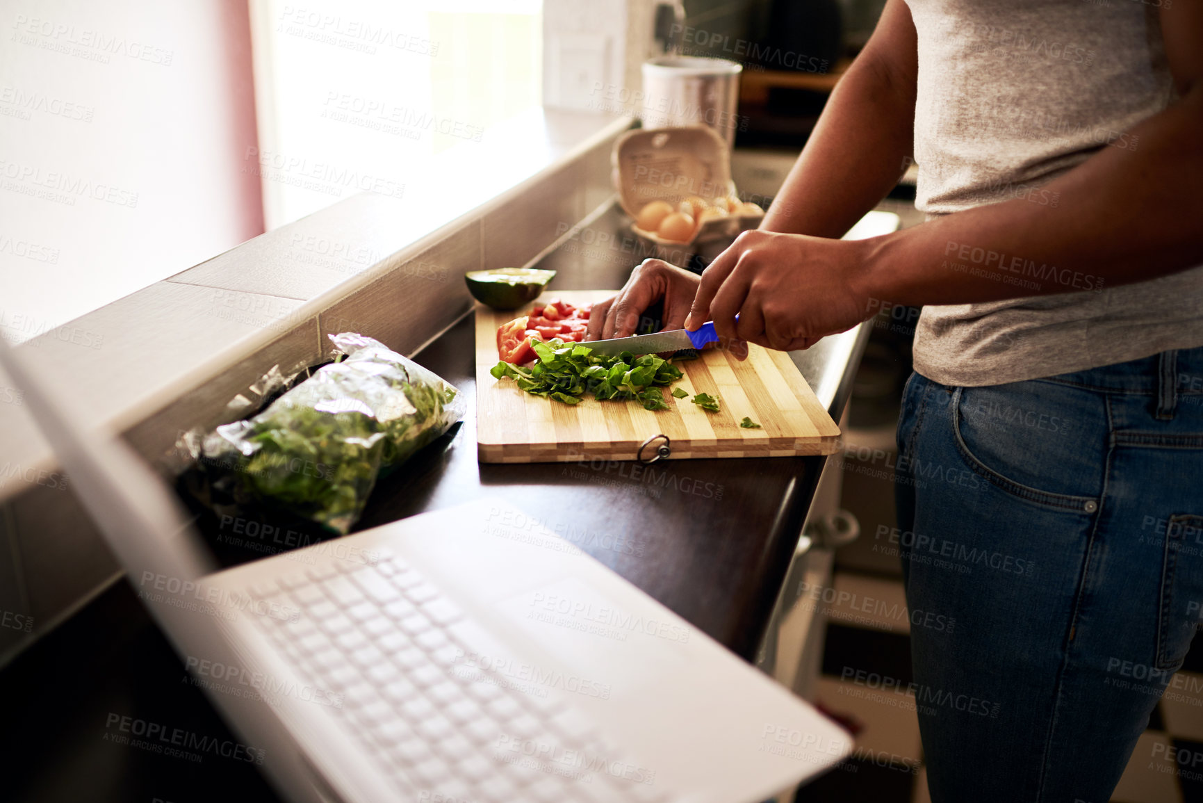 Buy stock photo Cropped shot of an unrecognizable man using a laptop while chopping vegetables in his kitchen at home