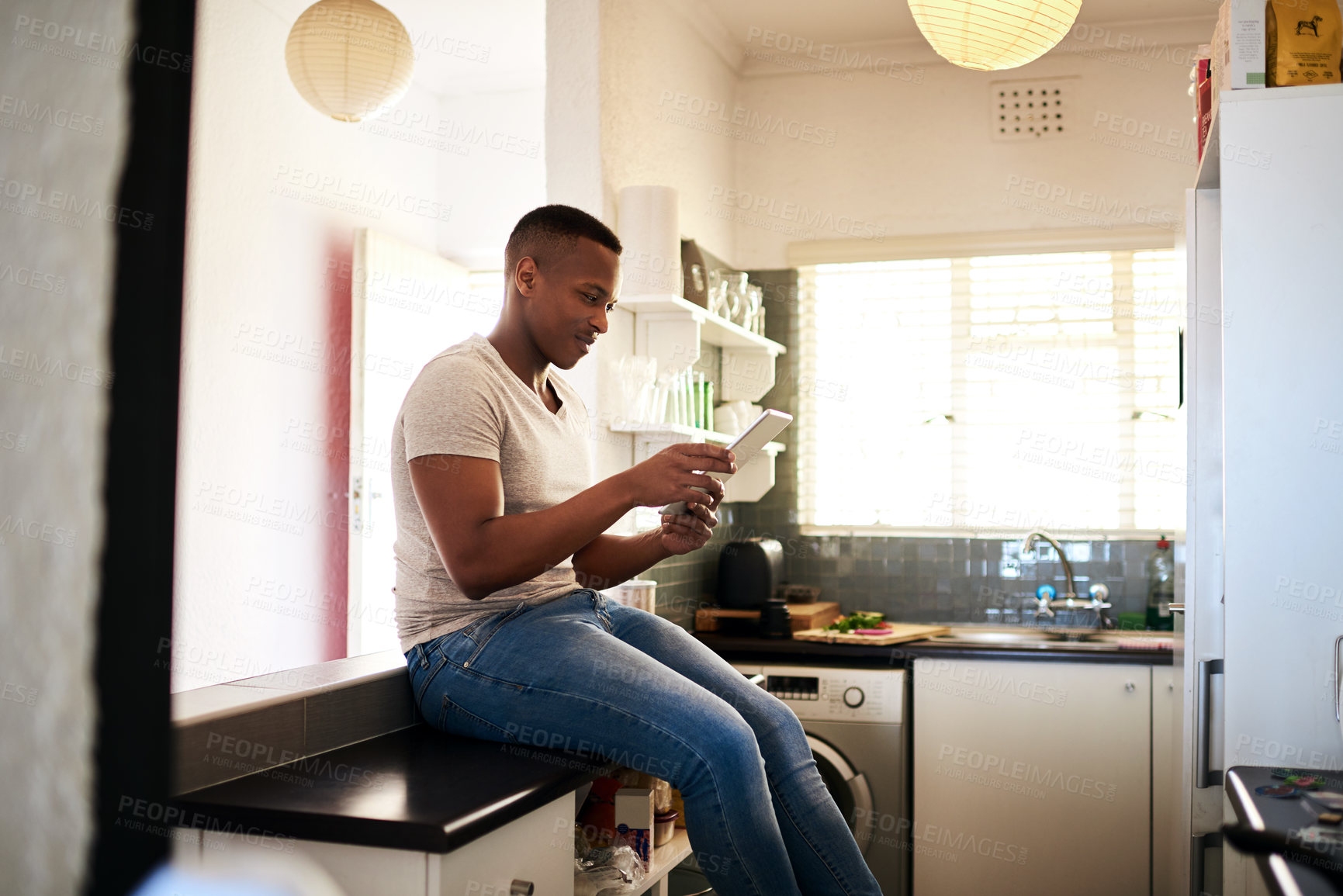Buy stock photo Cropped shot of a handsome young man using a digital tablet while sitting on the kitchen counter at home