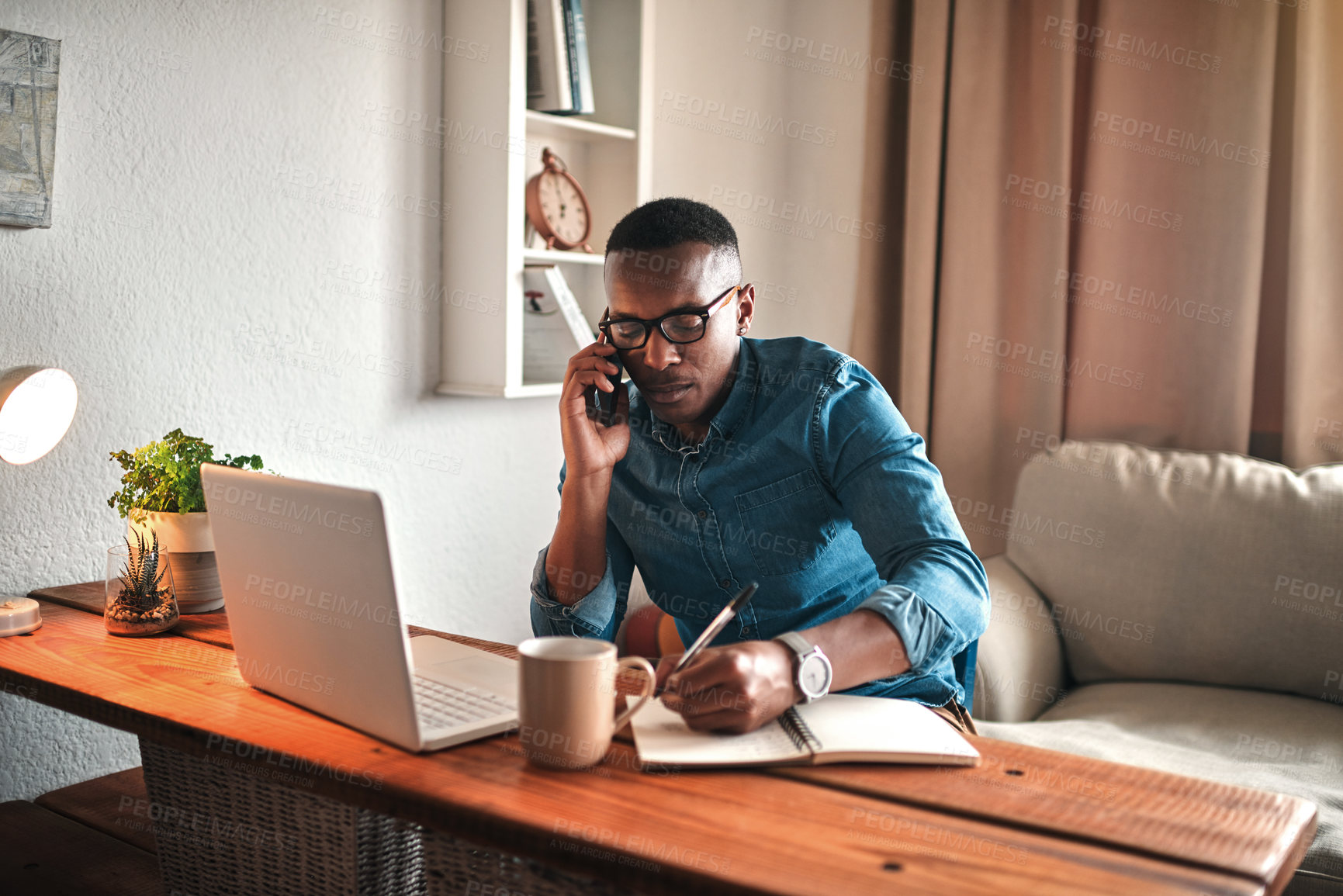 Buy stock photo Young entrepreneur on his phone while taking notes at his desk. Professional man working remote from home with technology. African American male has a business meeting on an audio call.