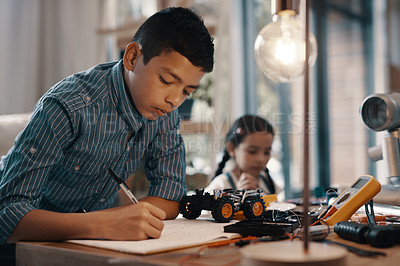 Buy stock photo Shot of a handsome young boy doing his homework on robotics at home with his younger sister in the background