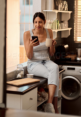 Buy stock photo Shot of a young woman using a smartphone while enjoying a cup of coffee at home
