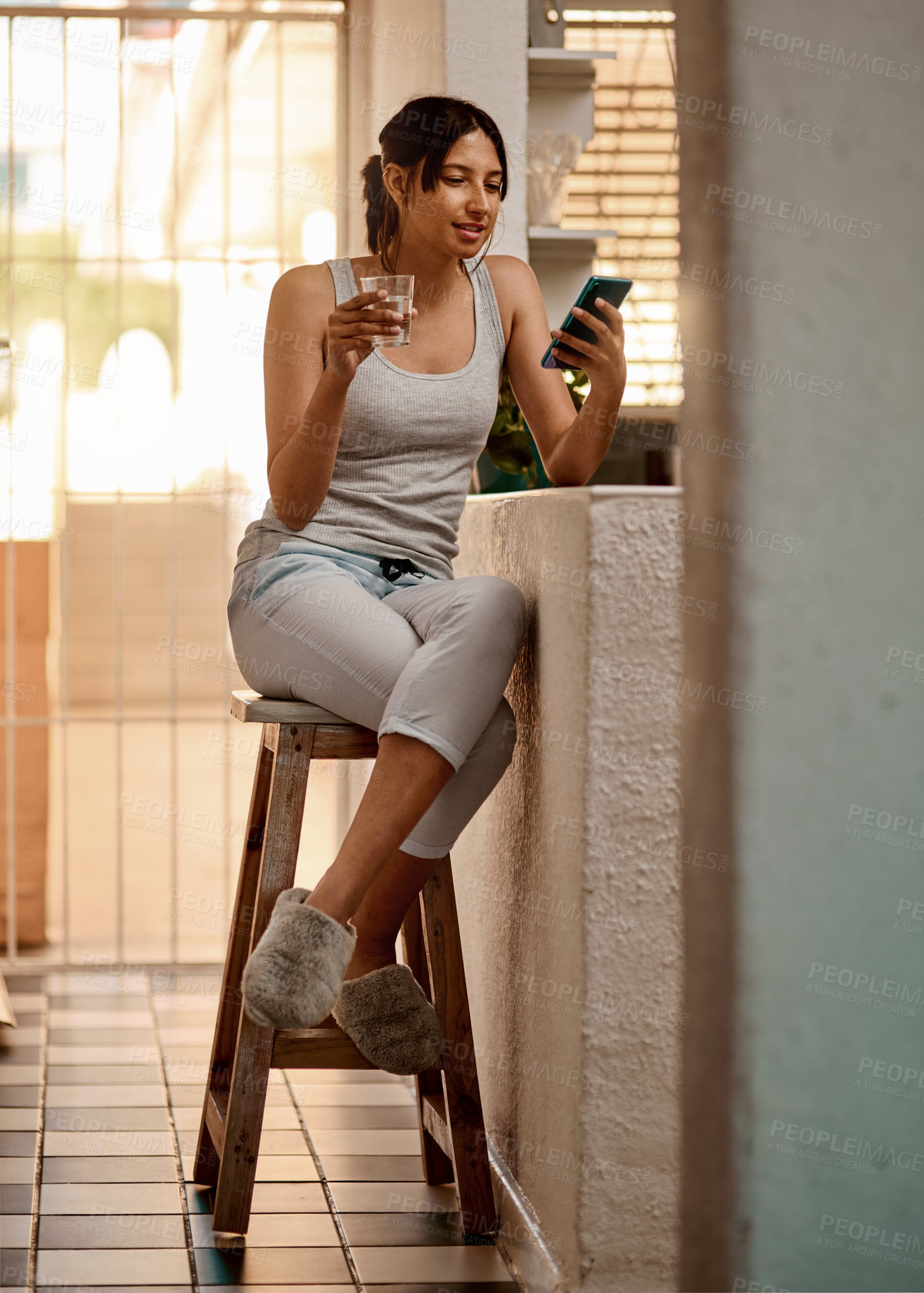 Buy stock photo Shot of a young woman using a smartphone and having a glass of water at home