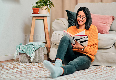 Buy stock photo Shot of a young woman relaxing on the floor in the living room and reading a book at home