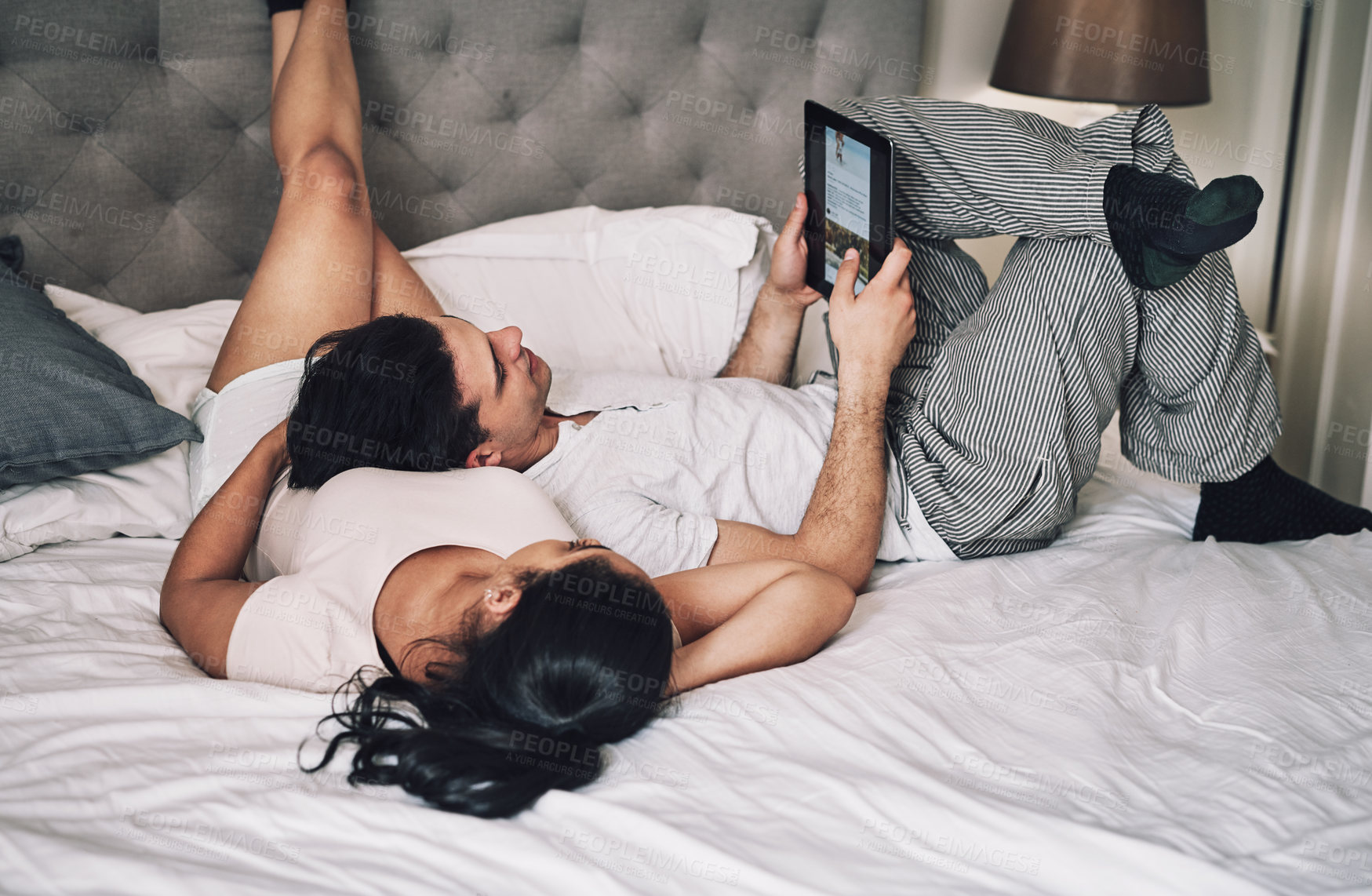 Buy stock photo Shot of a young couple looking at something on a digital tablet while lying in bed