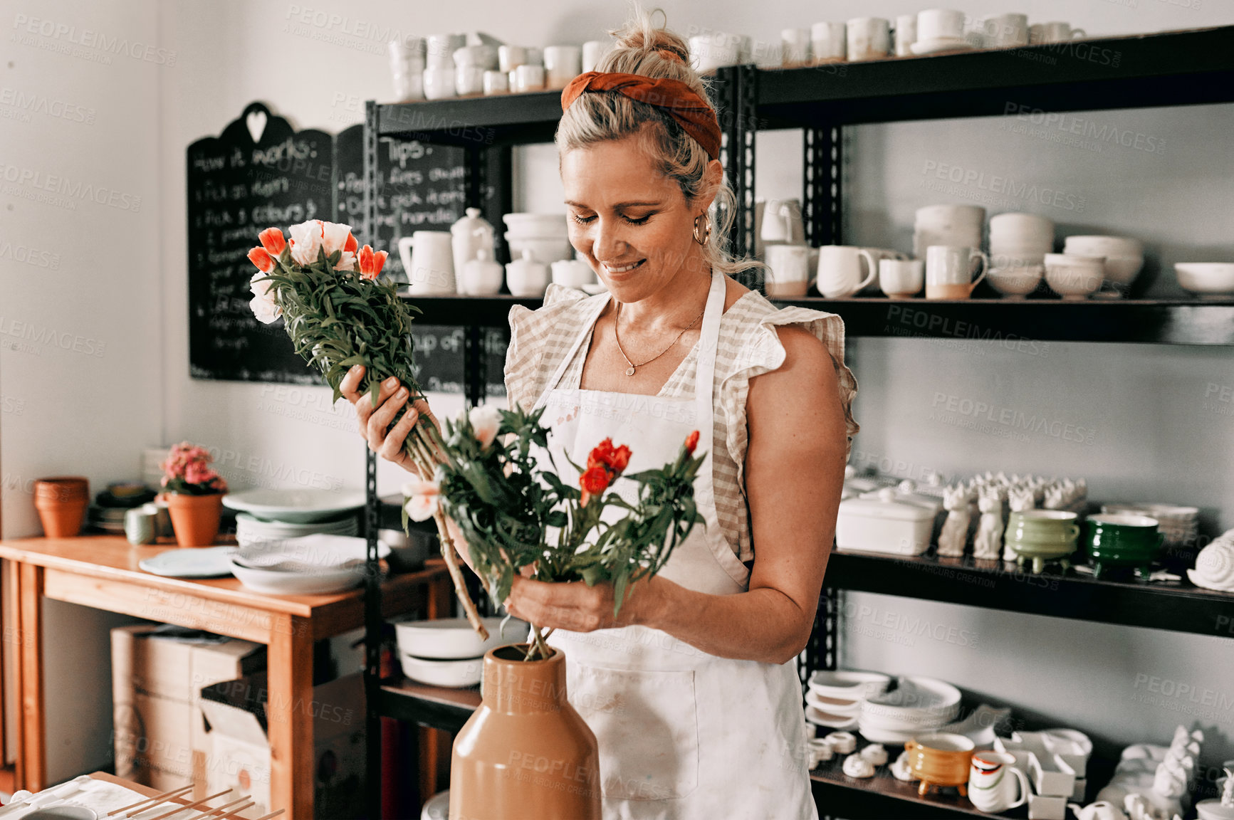 Buy stock photo Cropped shot of an attractive mature woman standing alone and putting flowers into a handmade vase in her pottery workshop