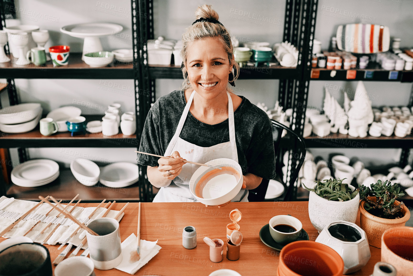 Buy stock photo Cropped portrait of an attractive mature woman sitting alone and painting a pottery bowl in her workshop