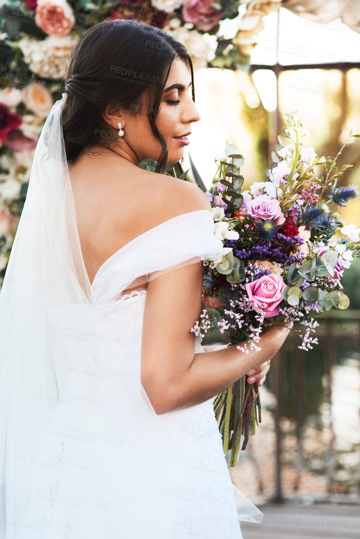 Buy stock photo Shot of a happy and beautiful young bride holding her bouquet of flowers while posing outdoors on her wedding day
