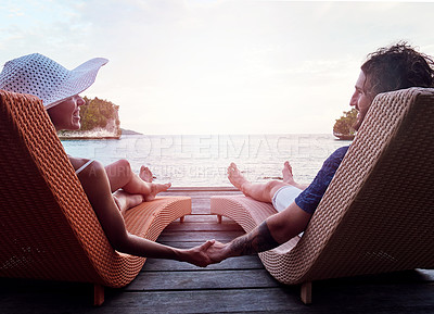 Buy stock photo Rearview shot of a cheerful young couple holding hands while relaxing on deck chairs and looking out at the ocean outside during the day